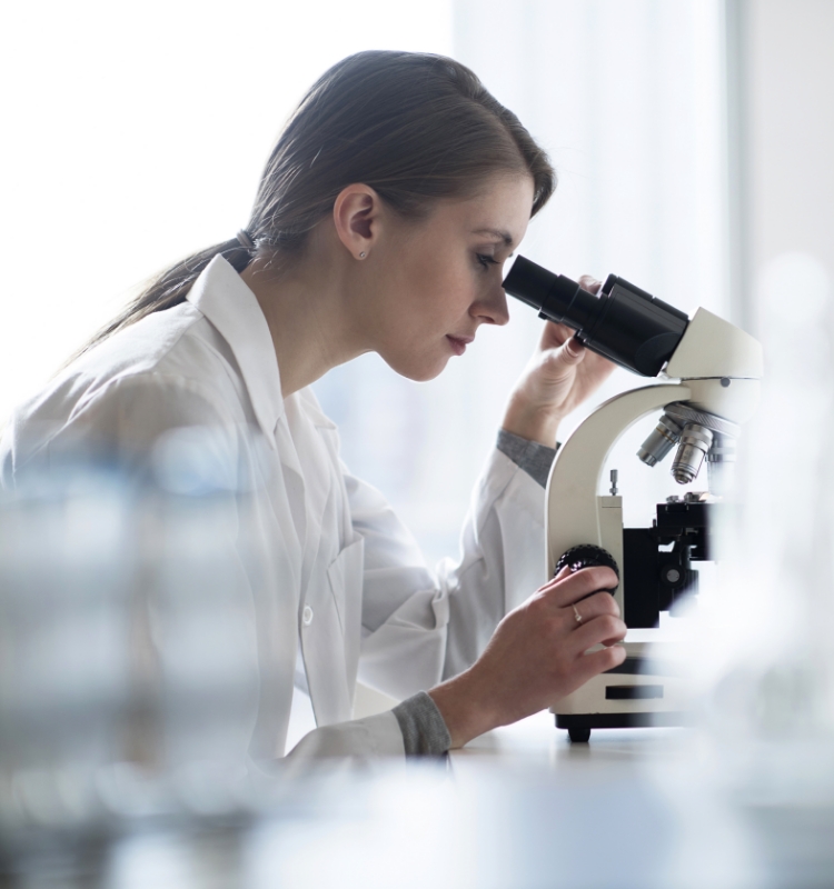 A scientist in a white lab coat examines a sample through a microscope in a bright laboratory.